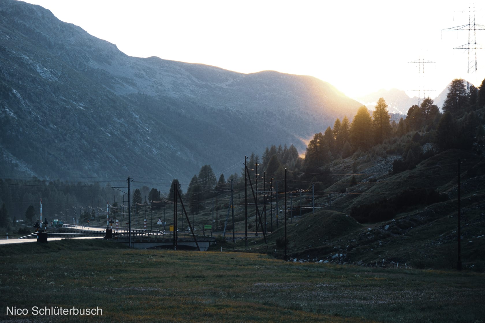 Sonnenuntergang über einem Alpental mit Passstraße und Bahntrasse