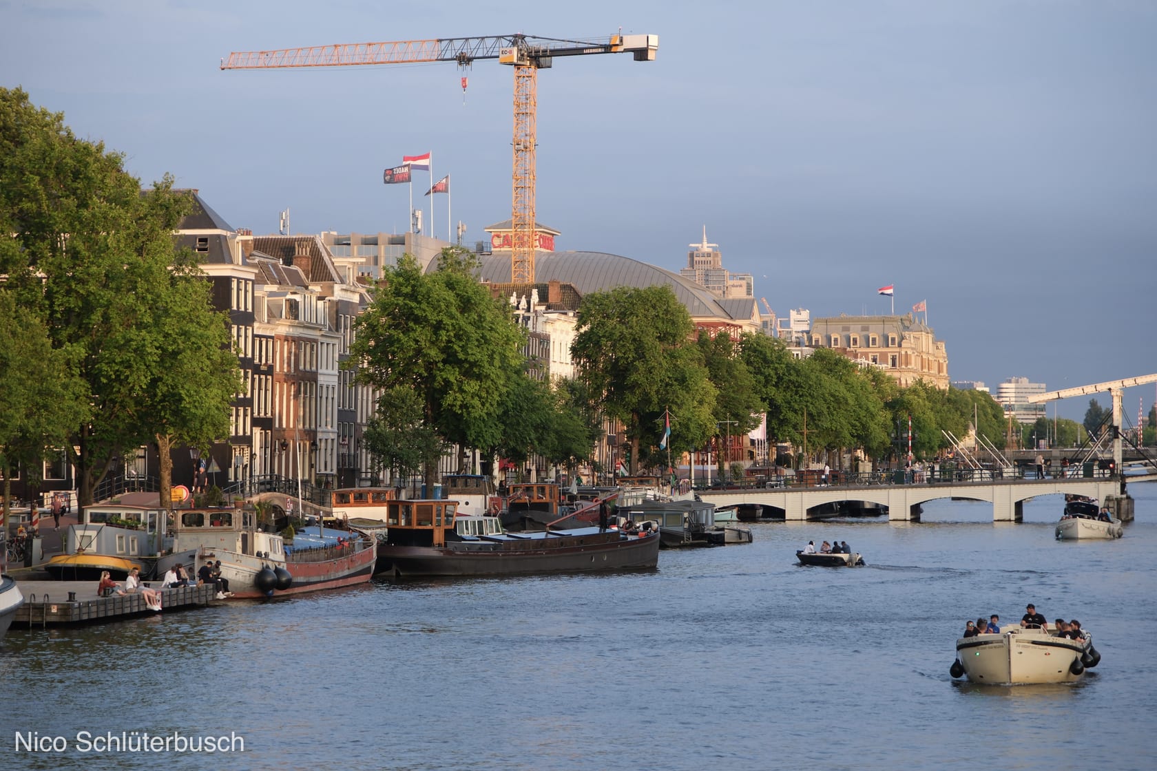 Abendlicht an der Amstel in Amsterdam mit vertäuten Hausbooten und Frachtkähnen am Kai, Ausflugsbooten auf dem Wasser und einem Baukran über der Häuserzeile