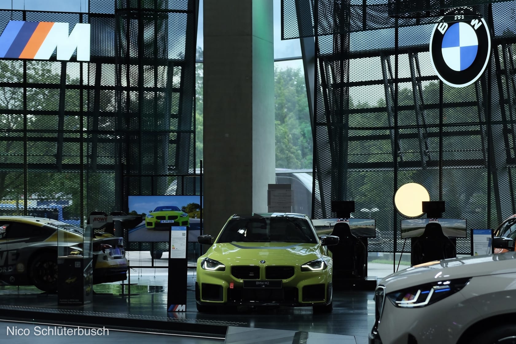 Gelbgrüner BMW M2 in der Frontansicht in einem Ausstellungsraum mit BMW- und M-Logo, umgeben von Rennsimulatoren und weiteren Fahrzeugen