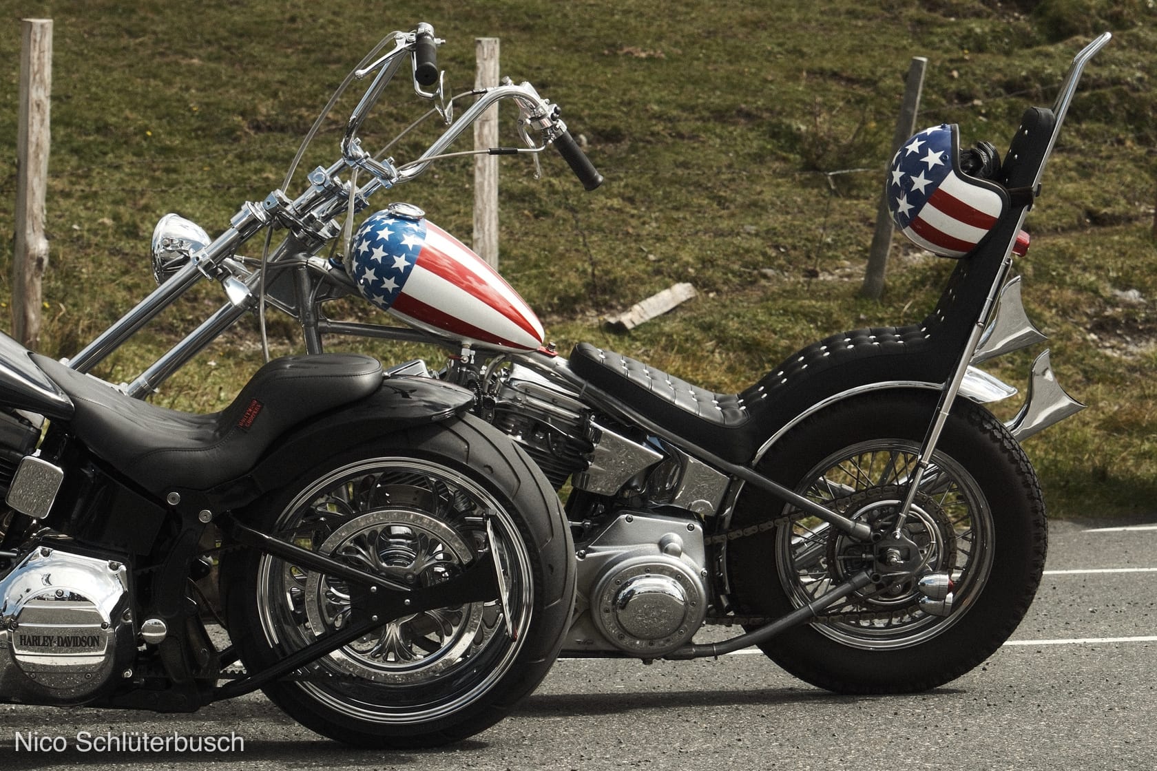 Harley-Davidson Chopper in der Seitenansicht mit Stars-and-Stripes-Tank, hoher Sissybar und passend lackiertem Helm vor einer Wiesenböschung