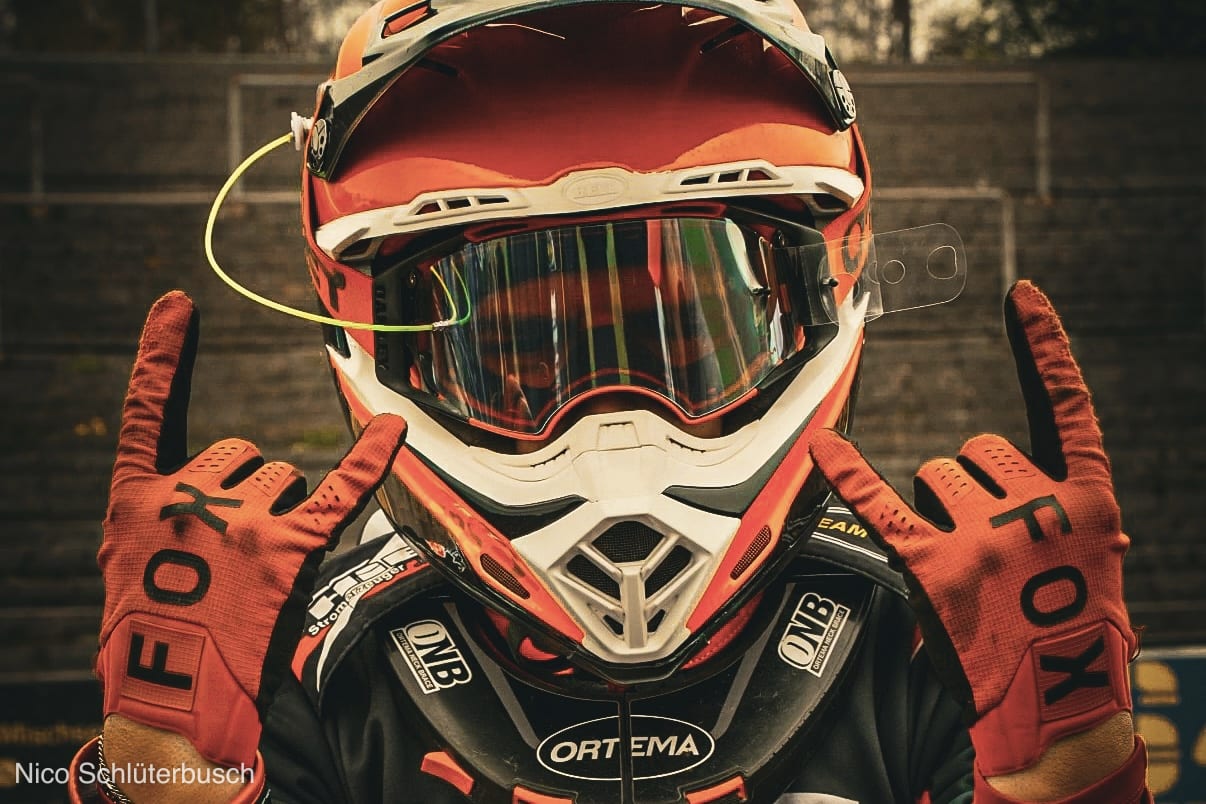 Motocross-Fahrer mit rot-weißem Helm, verspiegelter Brille und Nackenschutz zeigt mit beiden Fox-Handschuhen die Rock-Geste in die Kamera