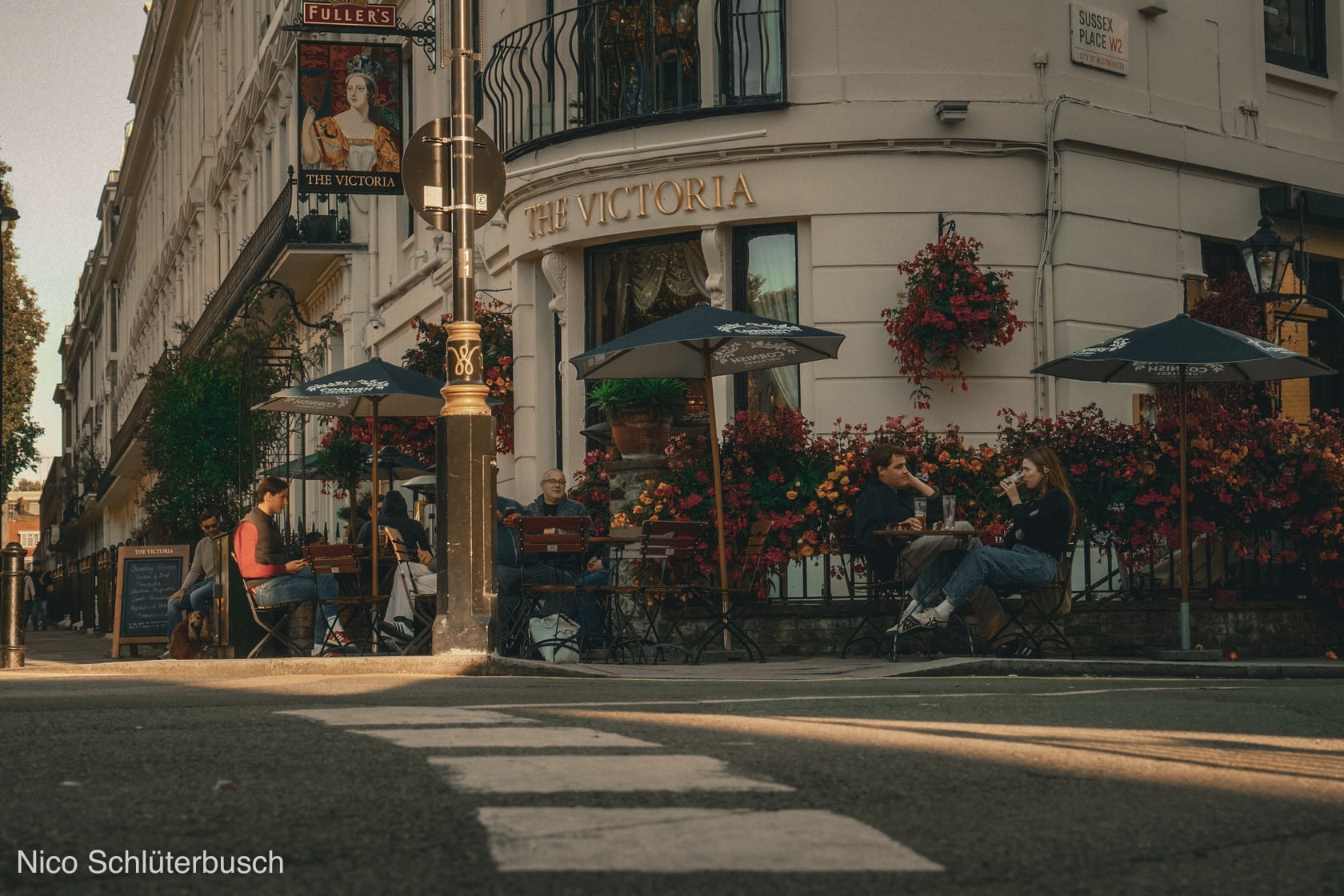 Straßenecke am Londoner Pub The Victoria mit blumengeschmückter Fassade und Gästen unter Sonnenschirmen in tiefstehender Sonne.