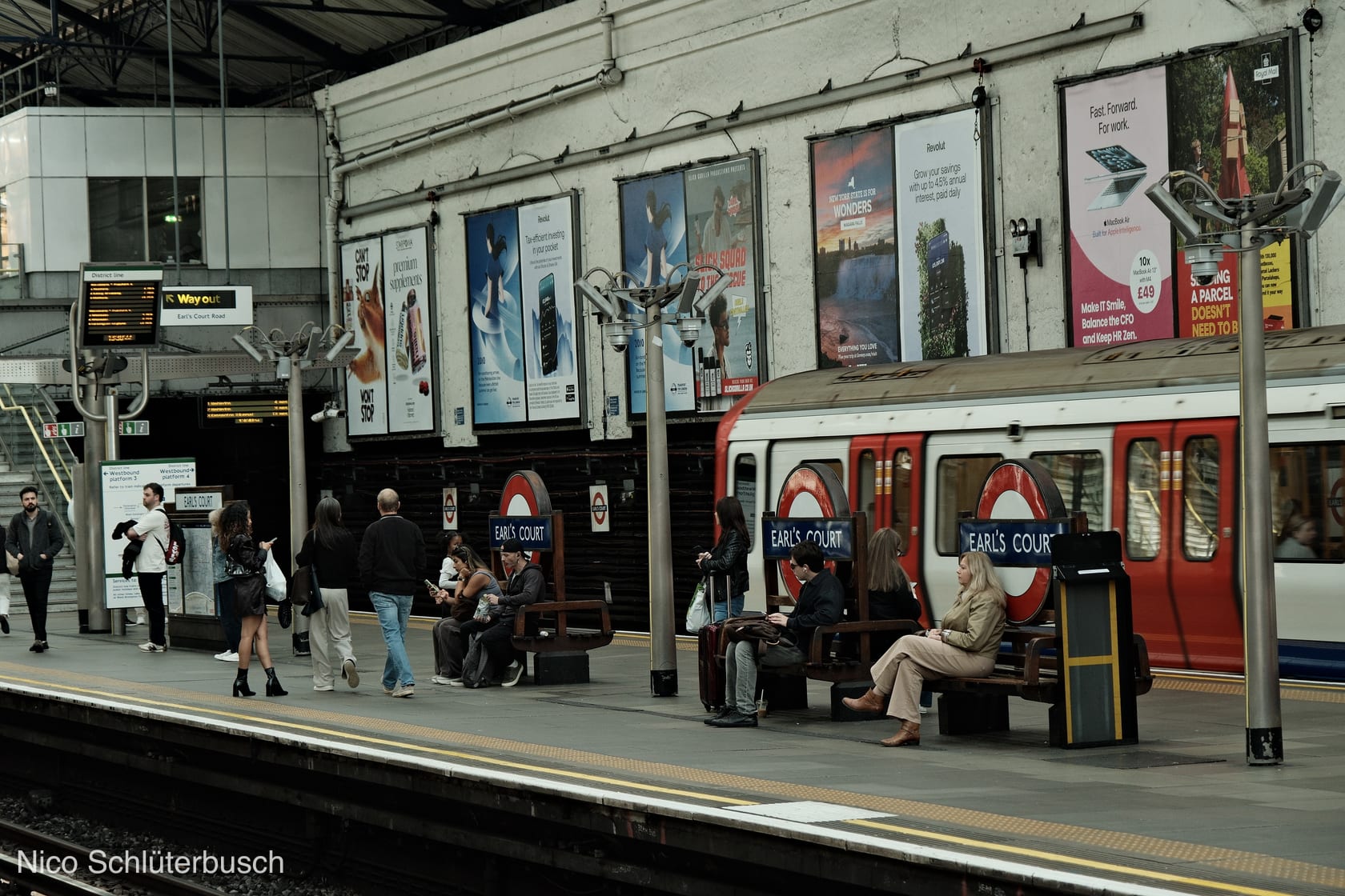 Wartende Fahrgäste auf dem Bahnsteig der Londoner U-Bahn-Station Earl's Court, rechts ein rot-weißer District-Line-Zug vor plakatierter Wand
