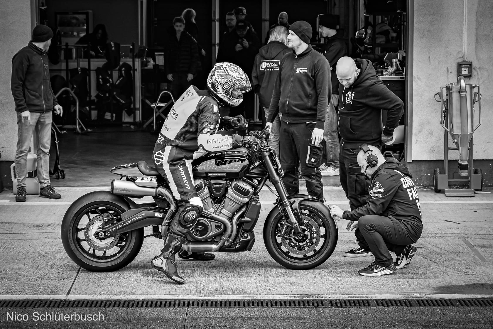 Rennfahrer in Lederkombi sitzt in der Boxengasse auf einer Harley-Davidson mit Slicks, während ein Mechaniker mit Headset kniend den Vorderreifen prüft