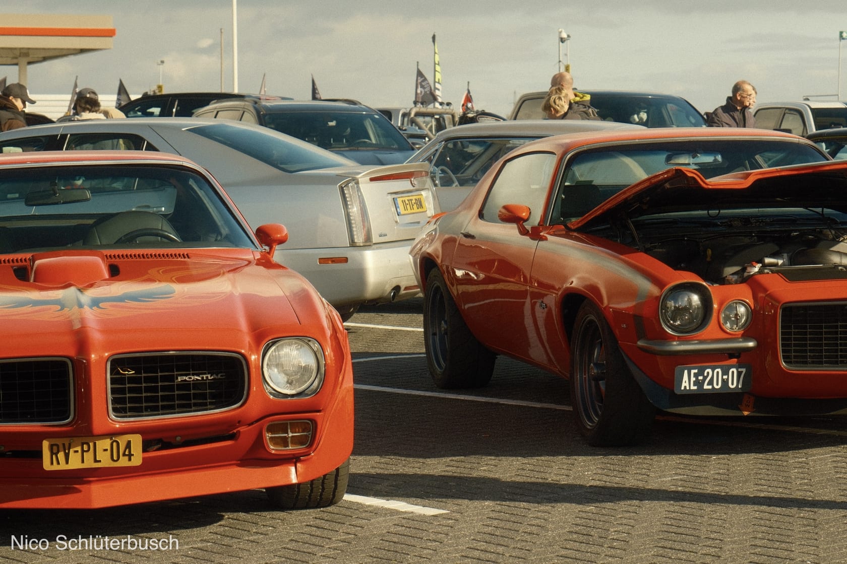 Orangeroter Pontiac Firebird und Chevrolet Camaro mit geöffneter Motorhaube nebeneinander auf einem gepflasterten Parkplatz bei einem US-Car-Treffen im Abendlicht.